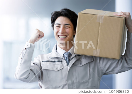 一個英俊的工人拿著一個紙箱,握著拳頭微笑。也有利於送貨和物流領域的工作和招聘。 一個英俊的工人拿著一個紙箱,握著拳頭微笑。也有利於送貨和物流領域的工作和招聘。 104986820