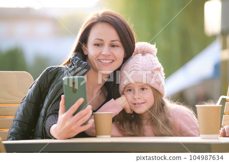 Young mother and her child daughter taking picture with phone selfie camera sitting at street cafe with hot drinks on sunny fall day. Social media presence in everyday life concept 104987634
