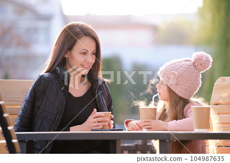 Young mother and her child daughter having good time together sitting at street cafe with hot drinks on sunny fall day. Happiness in family relations concept Young mother and her child daughter having good time together sitting at street cafe with hot drinks on sunny fall day. Happiness in family relations concept 104987635