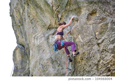 Strong female climber climbing steep wall of rocky mountain. Sportswoman overcoming difficult route. Engaging in extreme sports hobby concept. 104988004