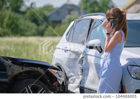 Stressed driver talking on sellphone on roadside near her smashed vehicle calling for emergency service help after car accident. Road safety and insurance concept 104988005