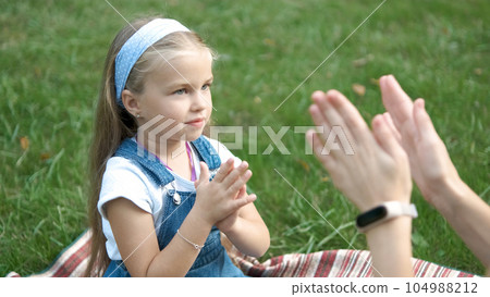 Pretty little child girl playing game with her mom clapping their hands together outdoors in green summer park. Pretty little child girl playing game with her mom clapping their hands together outdoors in green summer park. 104988212