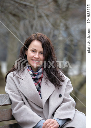 Portrait of happy pretty young woman with long dark hair resting on bench outdoors on spring sunny day 104988235