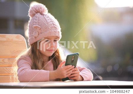 Portrait of cute little child girl in pink hat sitting alone at street cafe playing game on sellphone 104988247