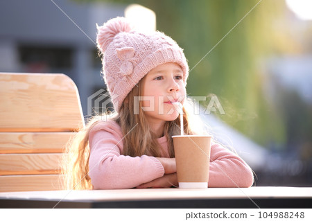 Portrait of cute little child girl in pink hat sitting alone at street cafe drinking tea from paper cup 104988248
