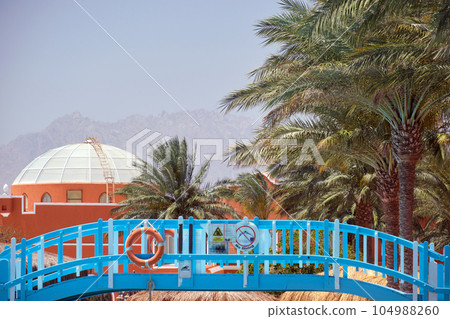 Picture from exotic summer holiday vacations. Pedestrian bridge to beach, building with round roof, palm trees and distant mountains 104988260