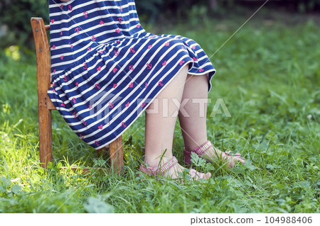 Legs and feet of little girl in dress sitting on a chair on green grass 104988406