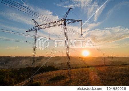 Dark silhouette of high voltage tower with electric power lines at sunrise. Transmission of electric energy concept Dark silhouette of high voltage tower with electric power lines at sunrise. Transmission of electric energy concept 104989026