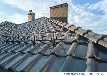 Closeup snow guard for safety in winter on house roof top covered with ceramic shingles. Tiled covering of building Closeup snow guard for safety in winter on house roof top covered with ceramic shingles. Tiled covering of building 104989122