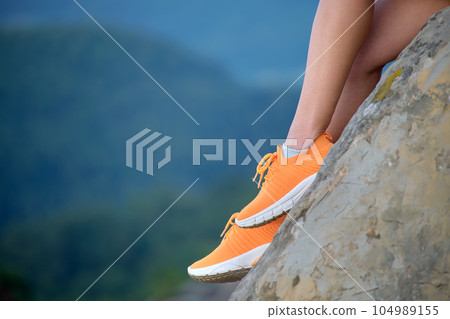 Closeup of woman hiker legs seated alone on rocky mountain cliff enjoying view of evening nature on wilderness trail. Active way of life concept 104989155