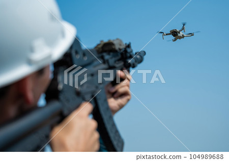 Caucasian man in a helmet shoots a flying drone with a rifle.  104989688