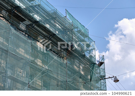 Building facade under renovation works with construction scaffolding frame covered with protective net for workers safety Building facade under renovation works with construction scaffolding frame covered with protective net for workers safety 104990621