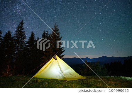 Brightly illuminated camping tent glowing on campsite in dark mountains under night stars covered sky. Active lifestyle and traveling concept. 104990625