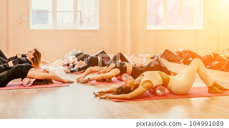 Happy middle aged well looking women, performing fascia exercises on floor using massage foam roller - tool to relieve tension in the back and relieve muscle pain. Female fitness yoga routine concept 104991089