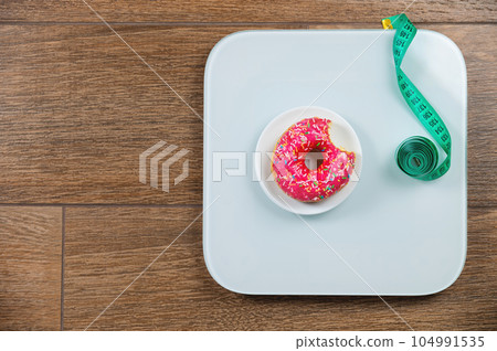 Diet concept. Scales holding sweet donut and measuring tape, choice between healthy and unhealthy food. View from above 104991535