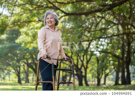 Happy old elderly Asian woman uses a walker for osteoarthritis rehabilitation physiotherapy in park.  Concept of happy retirement With care from a caregiver and Savings and senior health insurance 104994891