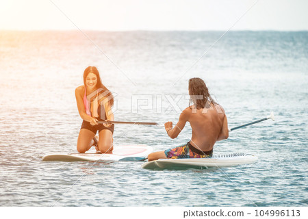 Sea woman and man on sup. Silhouette of happy young woman and man, surfing on SUP board, confident paddling through water surface. Idyllic sunset. Active lifestyle at sea or river. Sea woman and man on sup. Silhouette of happy young woman and man, surfing on SUP board, confident paddling through water surface. Idyllic sunset. Active lifestyle at sea or river. 104996113