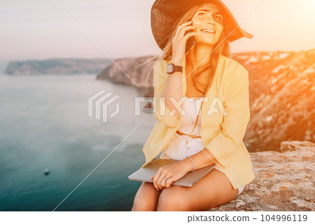 Digital nomad, Business woman working on laptop by the sea. Pretty lady typing on computer by the sea at sunset, makes a business transaction online from a distance. Freelance, remote work on vacation 104996119