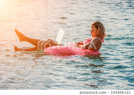 Woman works on laptop in sea. Freelancer, young blond woman in sunglases floating on an inflatable big pink donut with a laptop in the sea at sunset. Freelance, travel and holidays concept. 104996181