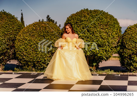 Woman yellow dress chess. A beautiful woman in a long puffy yellow dress poses on a chessboard in the park. Woman yellow dress chess. A beautiful woman in a long puffy yellow dress poses on a chessboard in the park. 104996265