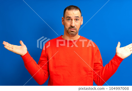 Bearded Hispanic man in his 40s wearing a red jumper raising his palms in a gesture of indifference, isolated on blue studio background. Bearded Hispanic man in his 40s wearing a red jumper raising his palms in a gesture of indifference, isolated on blue studio background. 104997008