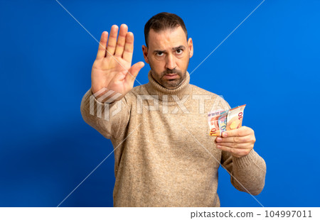Bearded hispanic man in his 40s wearing a beige turtleneck holding several euro bills and making the stop sign isolated over blue background Bearded hispanic man in his 40s wearing a beige turtleneck holding several euro bills and making the stop sign isolated over blue background 104997011