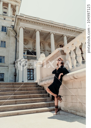 Stylish woman in the city. Fashion photo of a beautiful model in an elegant black dress posing against the backdrop of a building on a city street 104997125