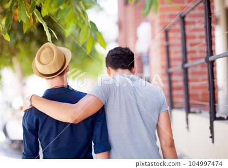 City, travel and back of gay couple walking together in the street while on a tourism vacation. Love, affection and male friends embracing in an outdoor urban road while on a weekend trip or holiday. 104997474
