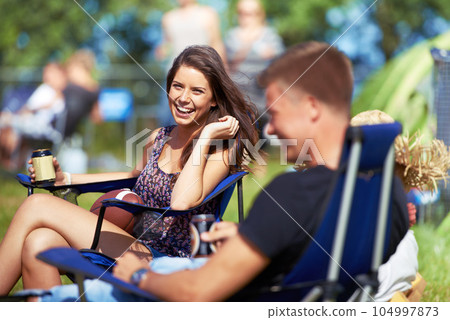 Portrait, woman drinking a beer at music festival or camping site and smiling with her friend. Vacation, happy female person sitting on camp chair and having a beverage while talking to her a male 104997873