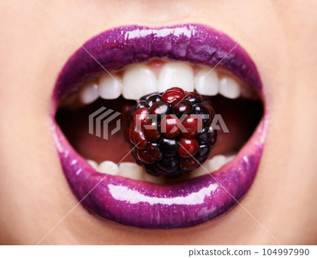 Closeup of woman lips with purple lipstick, makeup and beauty with shine and vibrant aesthetic, creativity and glow. Fruit between female model teeth, cosmetic product for mouth and cosmetology 104997990