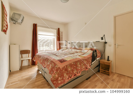 a bed in a room with wood flooring and red bedspreed sheets on the bed, next to a window 104999160