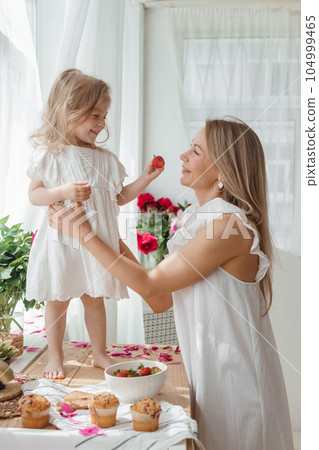 A little blonde girl with her mom on a kitchen countertop decorated with peonies. The concept of the relationship between mother and daughter. Spring atmosphere. A little blonde girl with her mom on a kitchen countertop decorated with peonies. The concept of the relationship between mother and daughter. Spring atmosphere. 104999465