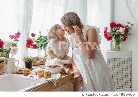 A little blonde girl with her mom on a kitchen countertop decorated with peonies. The concept of the relationship between mother and daughter. Spring atmosphere. A little blonde girl with her mom on a kitchen countertop decorated with peonies. The concept of the relationship between mother and daughter. Spring atmosphere. 104999469