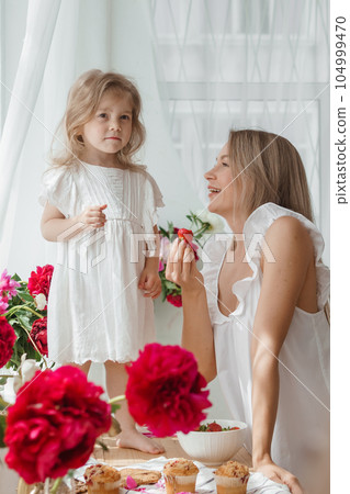A little blonde girl with her mom on a kitchen countertop decorated with peonies. The concept of the relationship between mother and daughter. Spring atmosphere. A little blonde girl with her mom on a kitchen countertop decorated with peonies. The concept of the relationship between mother and daughter. Spring atmosphere. 104999470