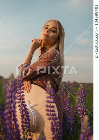 A beautiful woman in a straw hat walks in a field with purple flowers. A walk in nature in the lupin field A beautiful woman in a straw hat walks in a field with purple flowers. A walk in nature in the lupin field 104999481