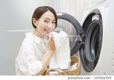 Smiling middle-aged woman smelling the laundry in front of the washing machine 105002593