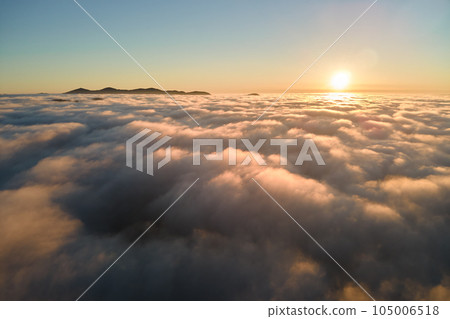 Aerial view of colorful sunrise over white dense fog with distant dark silhouettes of mountain hills on horizon Aerial view of colorful sunrise over white dense fog with distant dark silhouettes of mountain hills on horizon 105006518