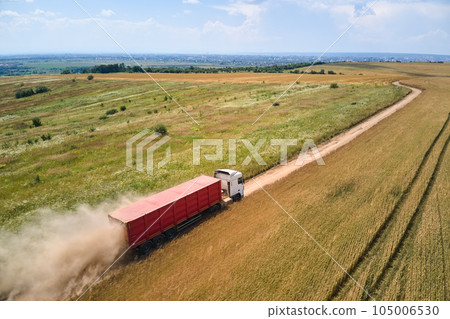 Aerial view of cargo truck driving on dirt road between agricultural wheat fields making lot of dust. Transportation of grain after being harvested by combine harvester during harvesting season Aerial view of cargo truck driving on dirt road between agricultural wheat fields making lot of dust. Transportation of grain after being harvested by combine harvester during harvesting season 105006530