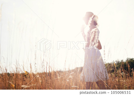 Portrait of young woman walking among high grasses in summer meadow wearing straw hat and linen dress enjoying nature. Harmony and balance Portrait of young woman walking among high grasses in summer meadow wearing straw hat and linen dress enjoying nature. Harmony and balance 105006531