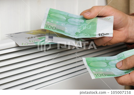 Human Hand Holding Euro Banknotes near Electric Heater Radiator at Home. Man Pay Money for Heating. Expensive to Heat. Energy Saving. Rising Cost of Living. Global Gas Crisis. Paying Cold Season Bills Human Hand Holding Euro Banknotes near Electric Heater Radiator at Home. Man Pay Money for Heating. Expensive to Heat. Energy Saving. Rising Cost of Living. Global Gas Crisis. Paying Cold Season Bills 105007158