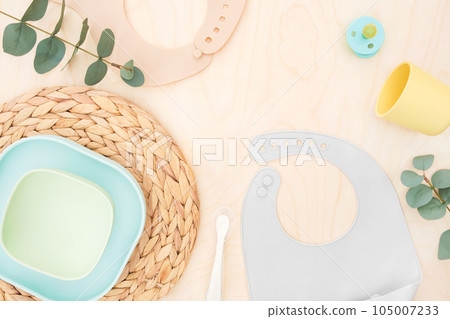 Baby accessories and tableware for food on wooden background. First feeding for kids concept. Flat lay, Top View, Space for text. Silicone dishware, bib, cup on neutral backdrop. Serving baby food 105007233