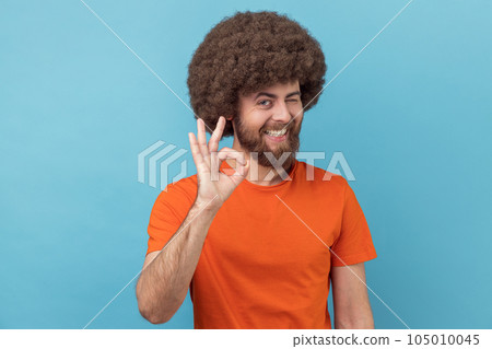 Portrait of positive man with Afro hairstyle wearing orange T-shirt standing and looking at camera with Ok sign, winking and toothy smile. Indoor studio shot isolated on blue background. 105010045