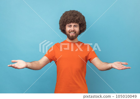 Man with Afro hairstyle wearing orange T-shirt sharing opening hands looking at camera with kind smile, greeting and regaling, happy glad to see you. Indoor studio shot isolated on blue background. Man with Afro hairstyle wearing orange T-shirt sharing opening hands looking at camera with kind smile, greeting and regaling, happy glad to see you. Indoor studio shot isolated on blue background. 105010049