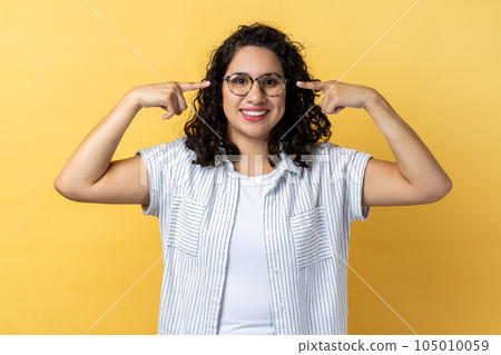 Portrait of smiling friendly positive woman with dark wavy hair in eyeglasses, standing pointing at her new spectacles, looking at camera. Indoor studio shot isolated on yellow background. Portrait of smiling friendly positive woman with dark wavy hair in eyeglasses, standing pointing at her new spectacles, looking at camera. Indoor studio shot isolated on yellow background. 105010059