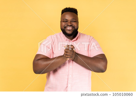 Portrait of happy satisfied friendly bearded man wearing pink shirt standing, keeping hands together, looking at camera with toothy smile. Indoor studio shot isolated on yellow background. 105010060