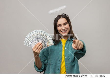 Happy angelic young woman with halo above head holding dollar banknotes and pointing to camera, choosing lottery winner, encouraging to earn big money. Indoor studio shot isolated on gray background. 105010070