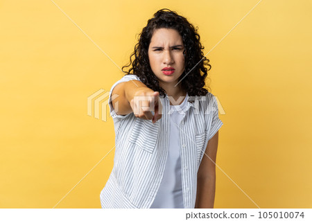 Portrait of strict woman with dark wavy hair noticing and pointing finger to camera, accusing with serious bossy face, making choice. Indoor studio shot isolated on yellow background. 105010074
