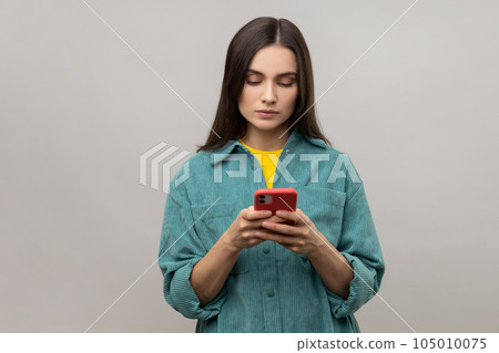 Serious pretty woman reading message on smartphone, using mobile device for communication, browsing web, wearing casual style jacket. Indoor studio shot isolated on gray background. 105010075