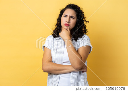 Portrait of pensive attractive woman with dark wavy hair being deeply in thoughts, keeping hand on head, looking away, holding chin. Indoor studio shot isolated on yellow background. Portrait of pensive attractive woman with dark wavy hair being deeply in thoughts, keeping hand on head, looking away, holding chin. Indoor studio shot isolated on yellow background. 105010076