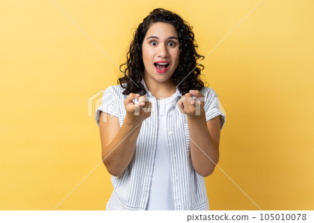 Portrait of amazed excited beautiful woman with dark wavy hair pointing directly at you, choosing something, has shocked expression. Indoor studio shot isolated on yellow background. 105010078